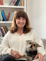 A woman sits on a couch with a dog on her lap, smiling.